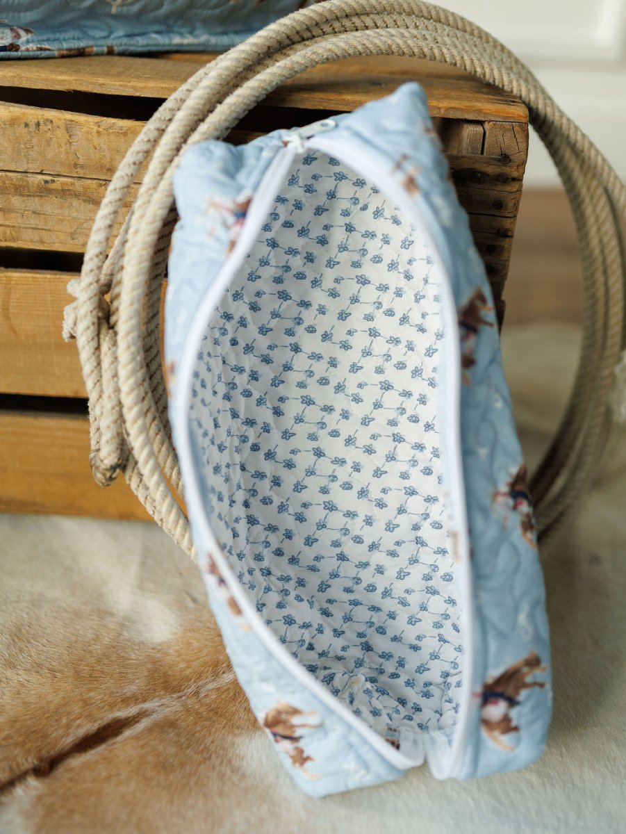 A Brumby Sky quilted bag set by Savannah Sevens western life{&}style, featuring a blue floral zippered pouch with white interior, rests on a wooden crate beside a coiled rope.