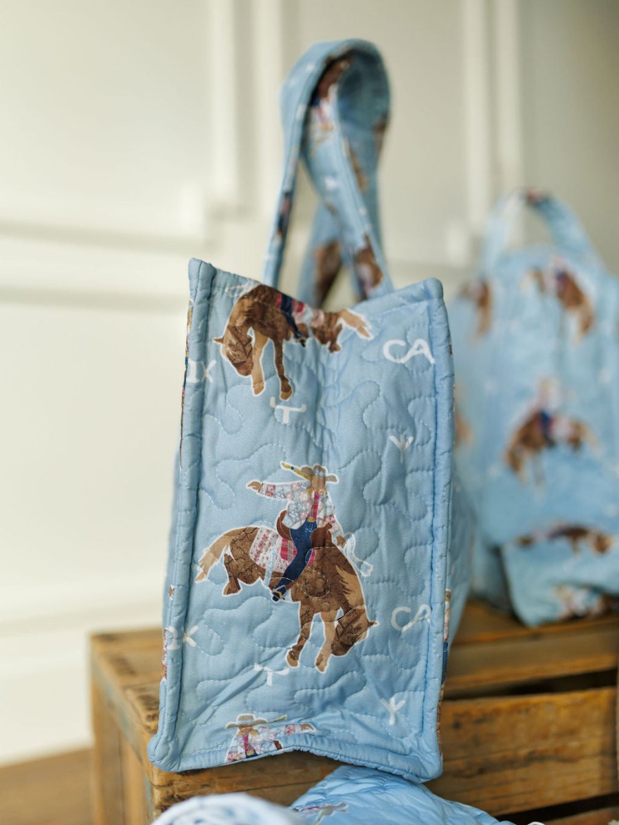 The Brumby Sky Collection tote by Savannah Sevens western life{&}style, in quilted light blue, showcases a rodeo cowgirl riding a bull. It’s displayed standing on a crate with matching quilted bags nearby against a softly blurred backdrop. By Ida Mae. 