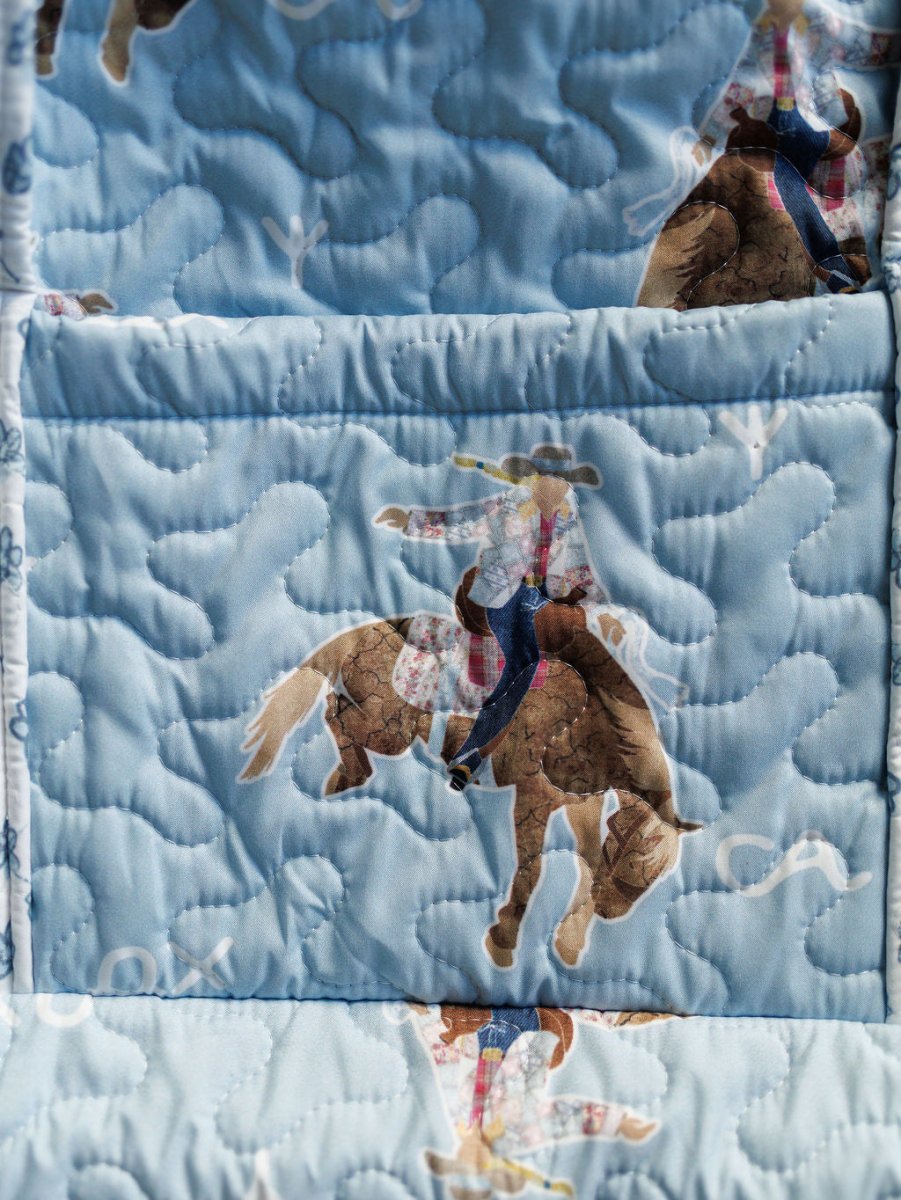 The Brumby Sky Collection by Savannah Sevens western life{&}style features a quilted bag set with light blue fabric adorned with cowboys in hats and plaid shirts riding brown horses, highlighted by swirling, cloud-like stitching. By Ida Mae. 