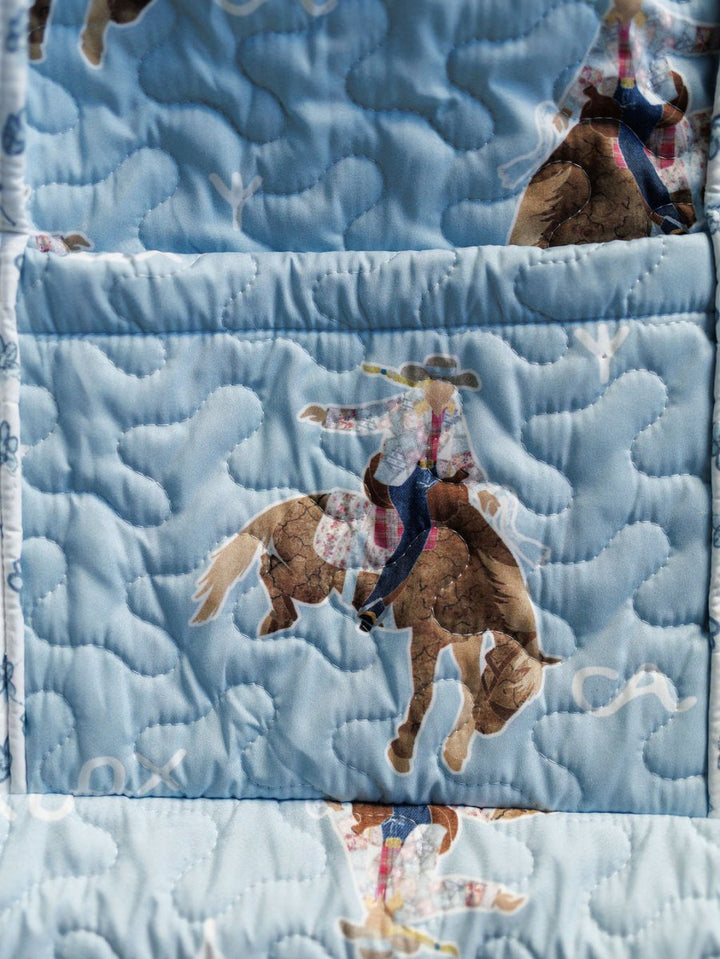 The Brumby Sky Collection by Savannah Sevens western life{&}style features a quilted bag set with light blue fabric adorned with cowboys in hats and plaid shirts riding brown horses, highlighted by swirling, cloud-like stitching. By Ida Mae. 
