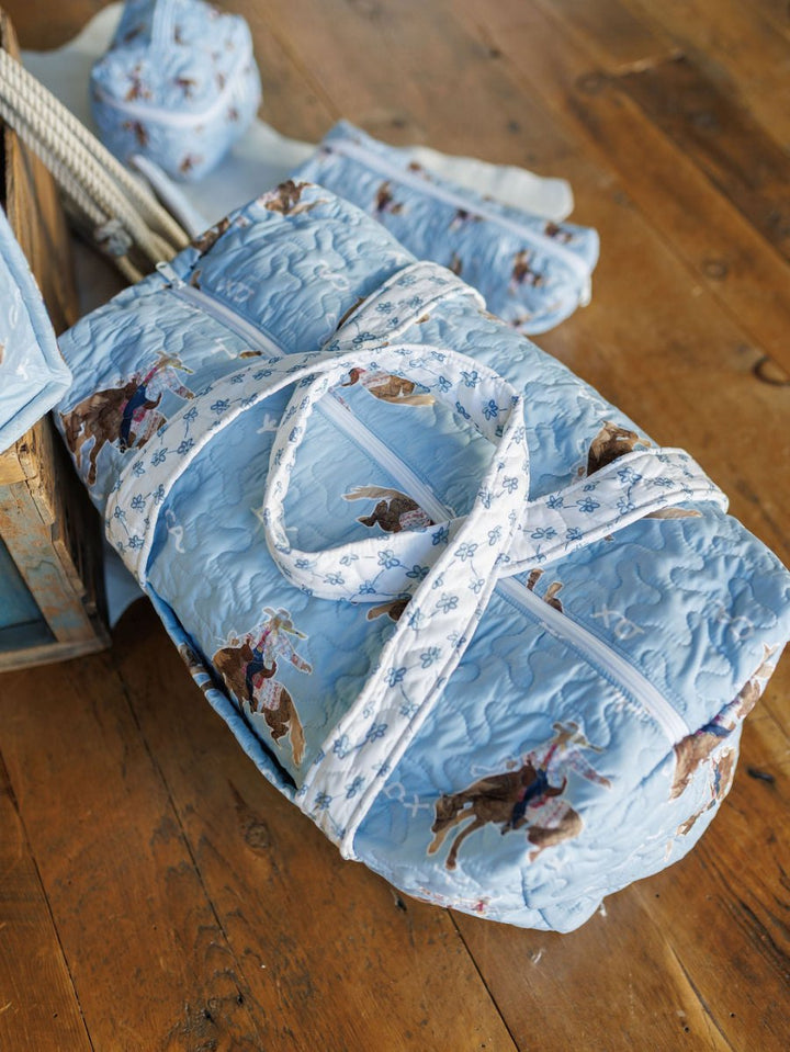 A quilted light blue duffel bag from the Brumby Sky Collection by Savannah Sevens western life{&}style, featuring a cowboy and horse print, sits on a wooden floor with matching set pieces in the background.