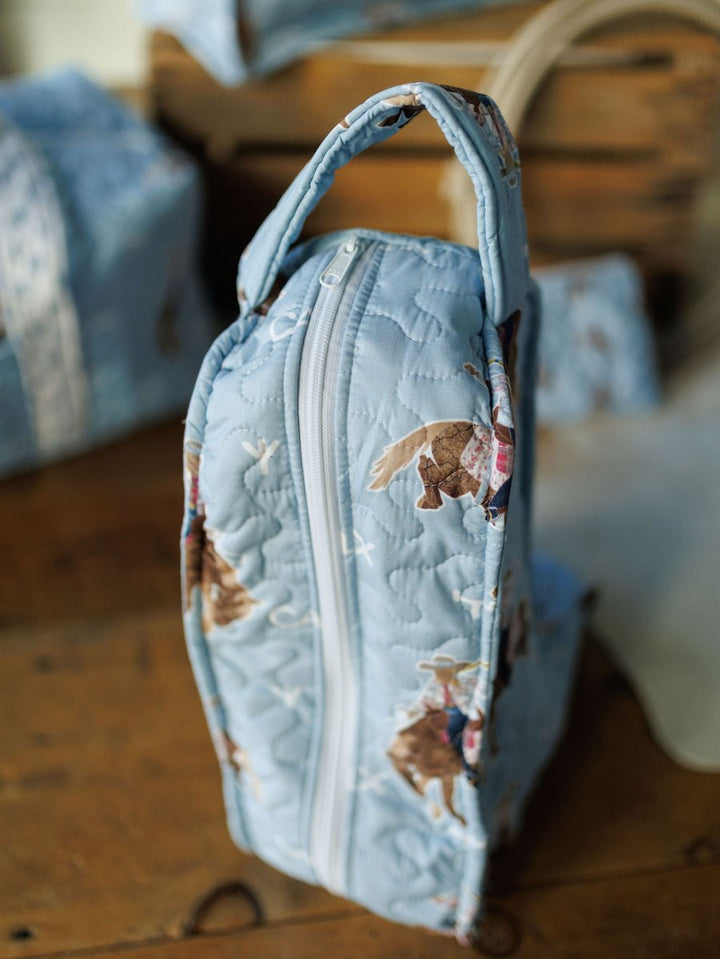 The Brumby Sky quilted bag by Savannah Sevens western life{&}style features a playful print of brown and white animals in red scarves and a white zipper. From the Brumby Sky Collection, it stands upright on a wooden surface. By Ida Mae. 