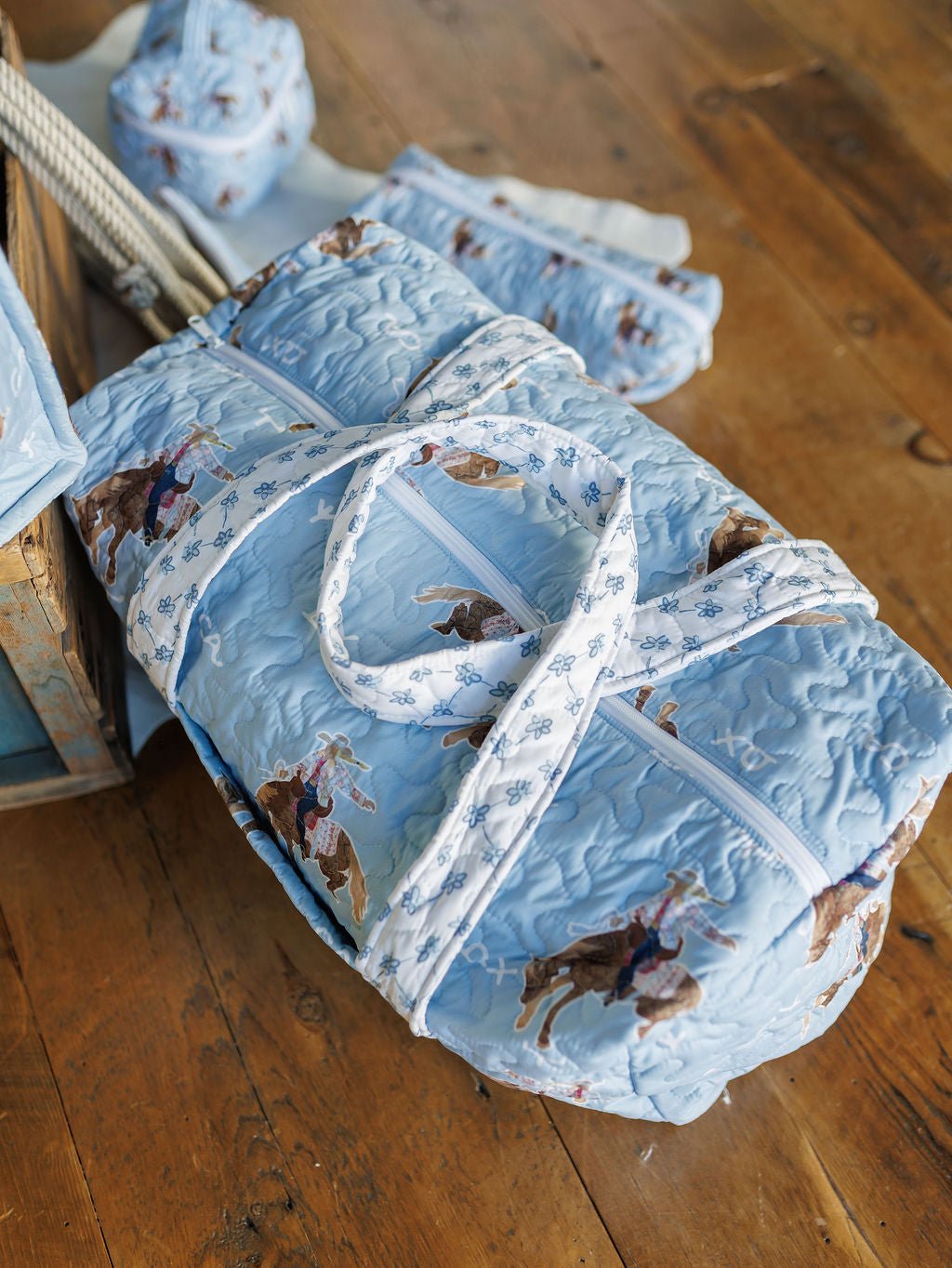 A quilted light blue duffel bag from the Brumby Sky Collection by Savannah Sevens western life{&}style, featuring a cowboy and horse print, sits on a wooden floor with matching set pieces in the background.