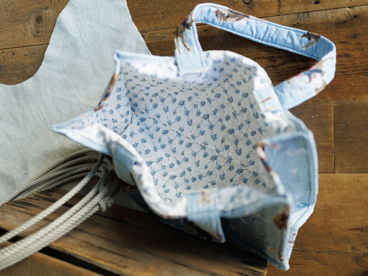 A quilted bag set from the Brumby Sky Collection by Savannah Sevens western life{&}style, featuring a fabric tote with light blue floral lining, shown from above on wood beside a white cloth with rope handles. By Ida Mae. 