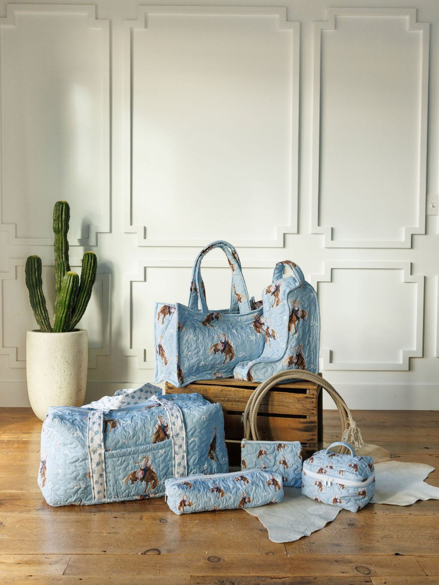 The Brumby Sky Collection by Savannah Sevens western life{&}style features a quilted light blue bag set with horse prints, styled on a wooden floor beside a cactus and crate, perfect for fans of western travel charm.