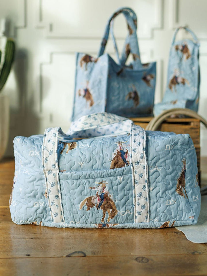The Brumby Sky Collection by Savannah Sevens western life{&}style—a light blue quilted duffel and tote with rodeo cowgirl and horse prints—rests on a wooden table in a sunlit room with white paneled walls.