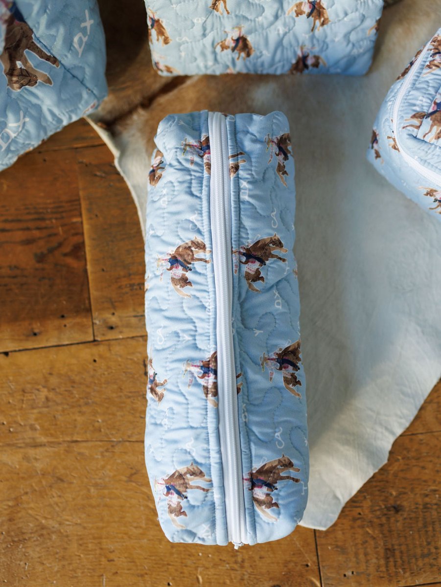 A quilted light blue zipper pouch from the Brumby Sky Collection by Savannah Sevens western life{&}style, featuring a cowboy and horse print, sits on a wooden surface beside similar quilted bag set items.