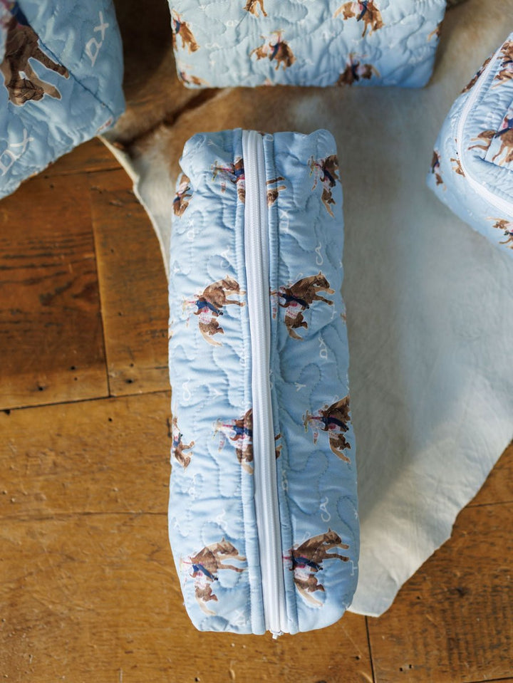 A quilted light blue zipper pouch from the Brumby Sky Collection by Savannah Sevens western life{&}style, featuring a cowboy and horse print, sits on a wooden surface beside similar quilted bag set items.