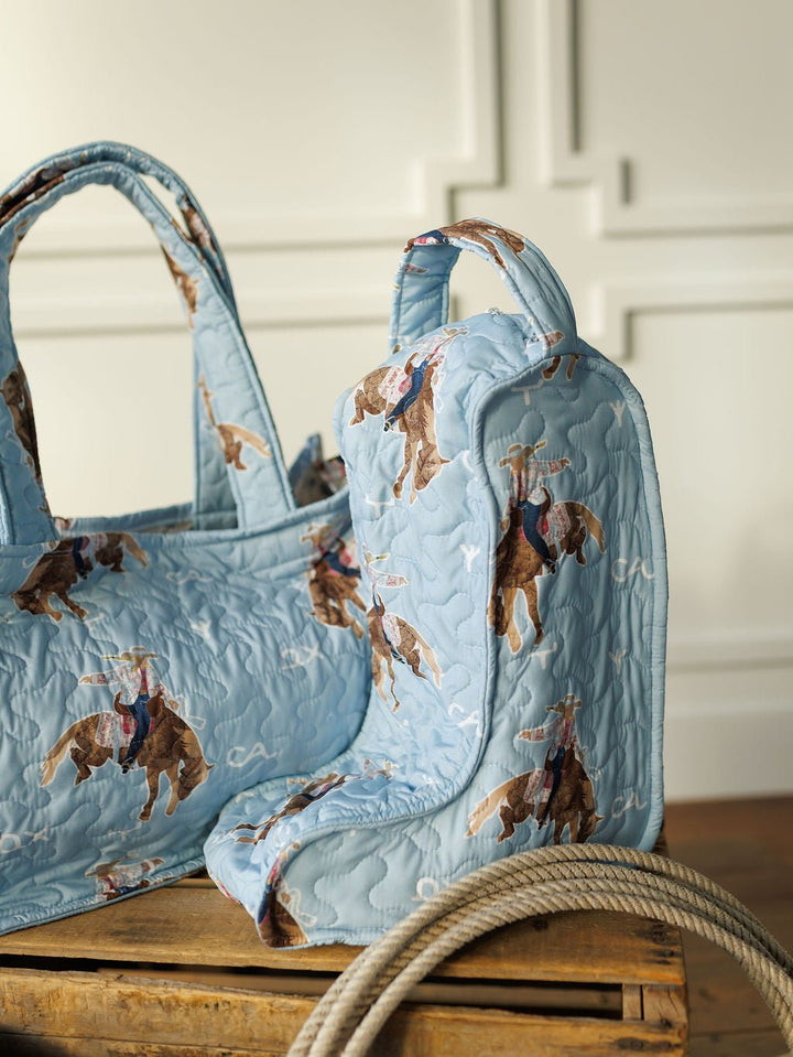 The Brumby Sky Collection quilted bag set and matching boot bag by Savannah Sevens western life&style, featuring a cowboy on a bucking bronco, rest on a wooden crate with a coiled rope against white paneled walls. By Ida Mae. 