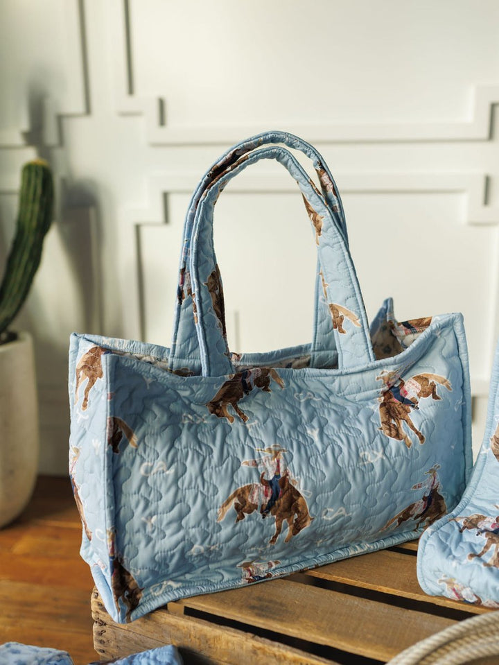 The Brumby Sky tote from Savannah Sevens western life&style features a light blue quilted design with playful cowboys on horseback, shown on wood with a white paneled wall and potted cactus in the background. By Ida Mae. 
