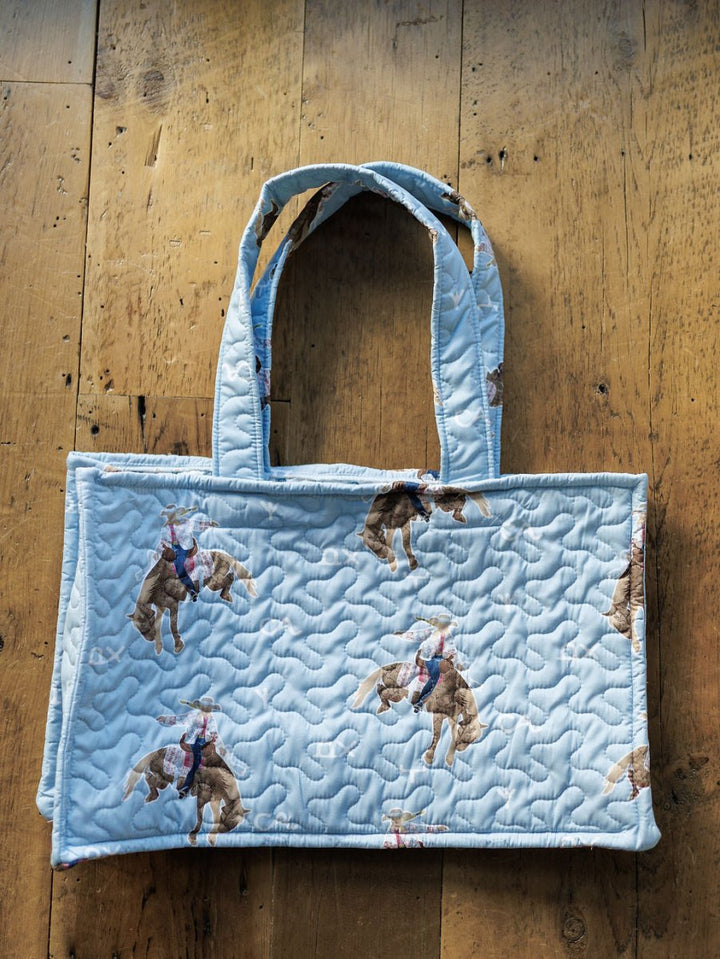 The Brumby Sky bag from Savannah Sevens western life&style features light blue quilted fabric, handles, and a repeating bucking horse pattern from the collection, shown on a wooden floor. By Ida Mae. 