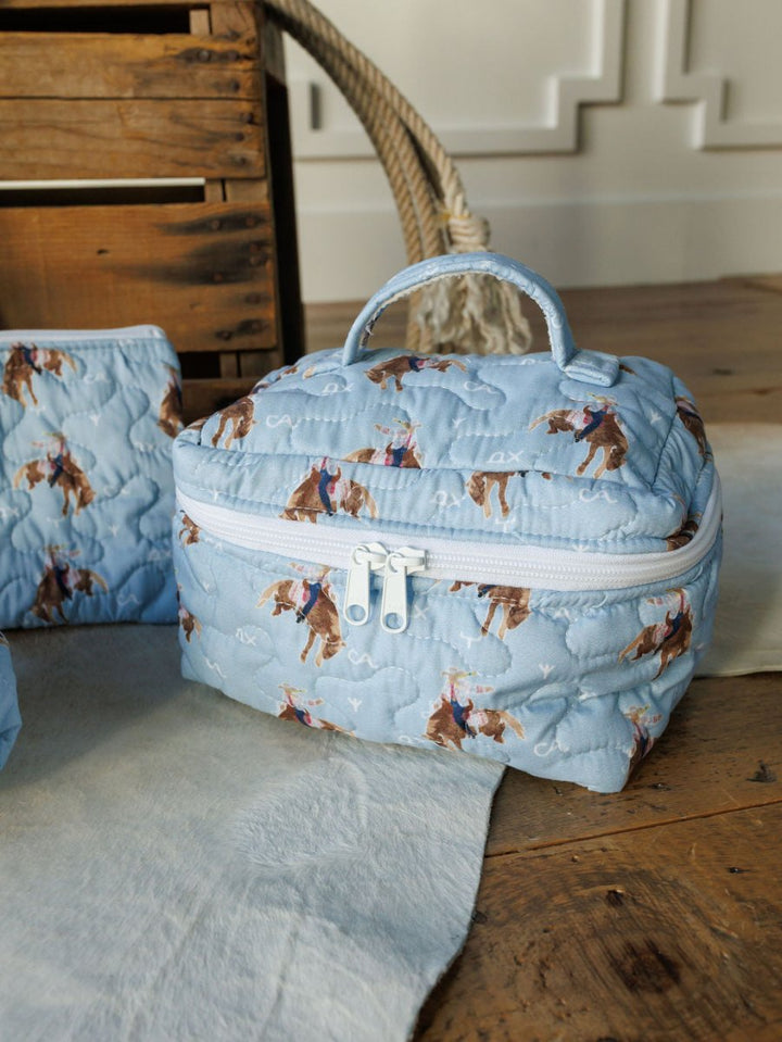 A quilted, light blue Brumby Sky cosmetic bag from Savannah Sevens western life{&}style features cowboy-and-horse motifs, a handle and zipper, and is shown on wood by a crate and braided basket handle. By Ida Mae.