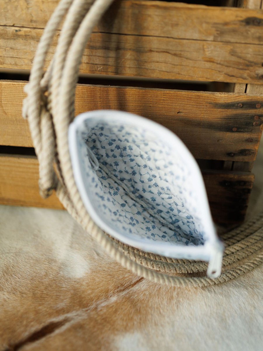 The Brumby Sky pouch from Savannah Sevens western life & style is open on a cowhide surface, showing its white and blue floral interior, beside a coiled rope and wooden crate. By Ida Mae. 