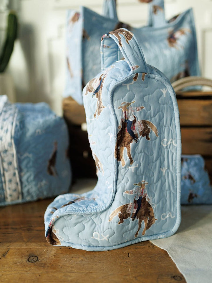 The Brumby Sky boot bag from Savannah Sevens western life{&}style’s collection features a cowboy and horse print in quilted light blue, shown with matching travel bags and fabric on a wooden table. By Ida Mae. 