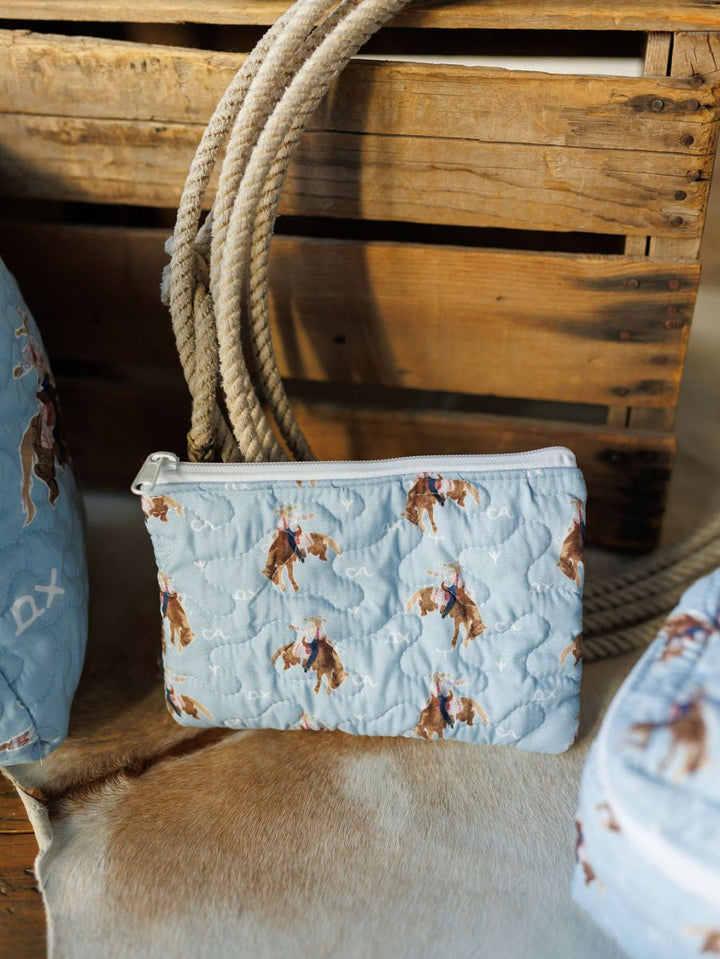 The Brumby Sky pouch from Savannah Sevens western life{&}style features a light blue quilted rodeo cowboy print with a white zipper, shown on brown and white cowhide before a wooden crate and coiled rope. By Ida Mae. 