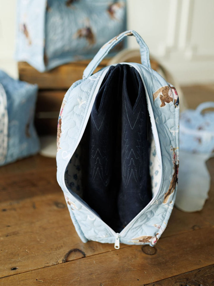 A pair of black cowboy boots are packed inside a Brumby Sky bag with a playful animal print from the Savannah Sevens western life&style Collection, sitting on a wooden floor. The light blue quilted bag features a zippered closure and handle. By Ida Mae. 