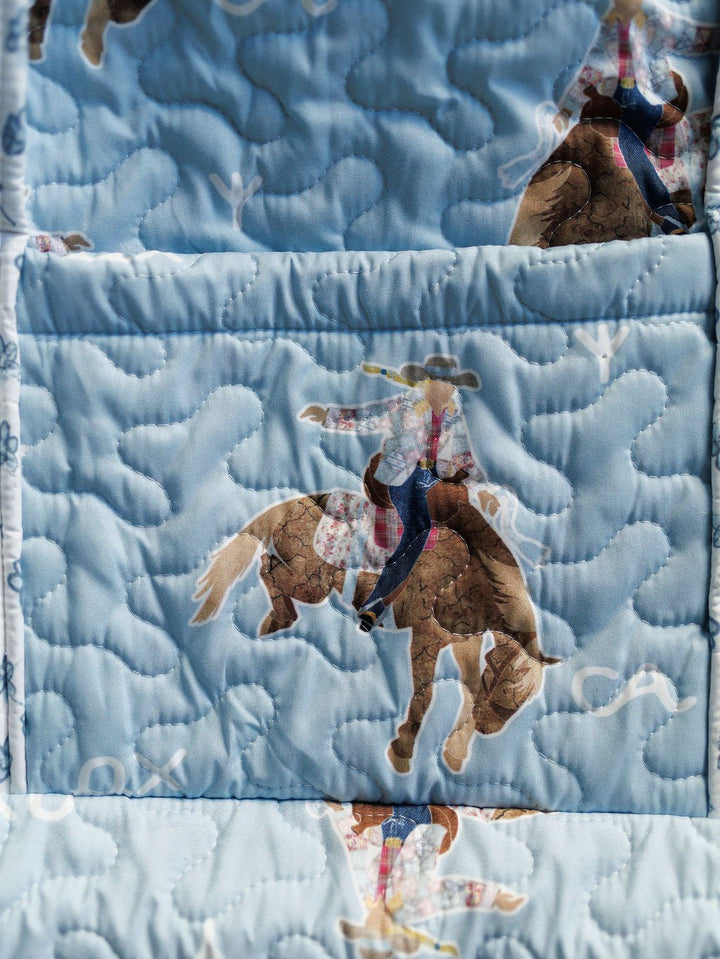 The Brumby Sky Collection by Savannah Sevens western life{&}style features a quilted bag set with light blue fabric adorned with cowboys in hats and plaid shirts riding brown horses, highlighted by swirling, cloud-like stitching. By Ida Mae. 