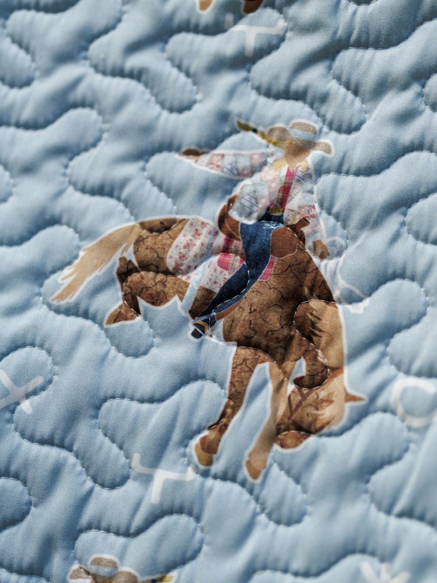 A close-up of quilted fabric from the Brumby Sky Collection by Savannah Sevens western life{&}style, featuring a cowboy on a bucking horse and blue wavy stitching over a light blue background. By Ida Mae. 