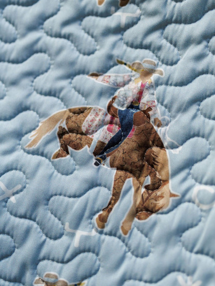A close-up of quilted fabric from the Brumby Sky Collection by Savannah Sevens western life{&}style, featuring a cowboy on a bucking horse and blue wavy stitching over a light blue background. By Ida Mae. 