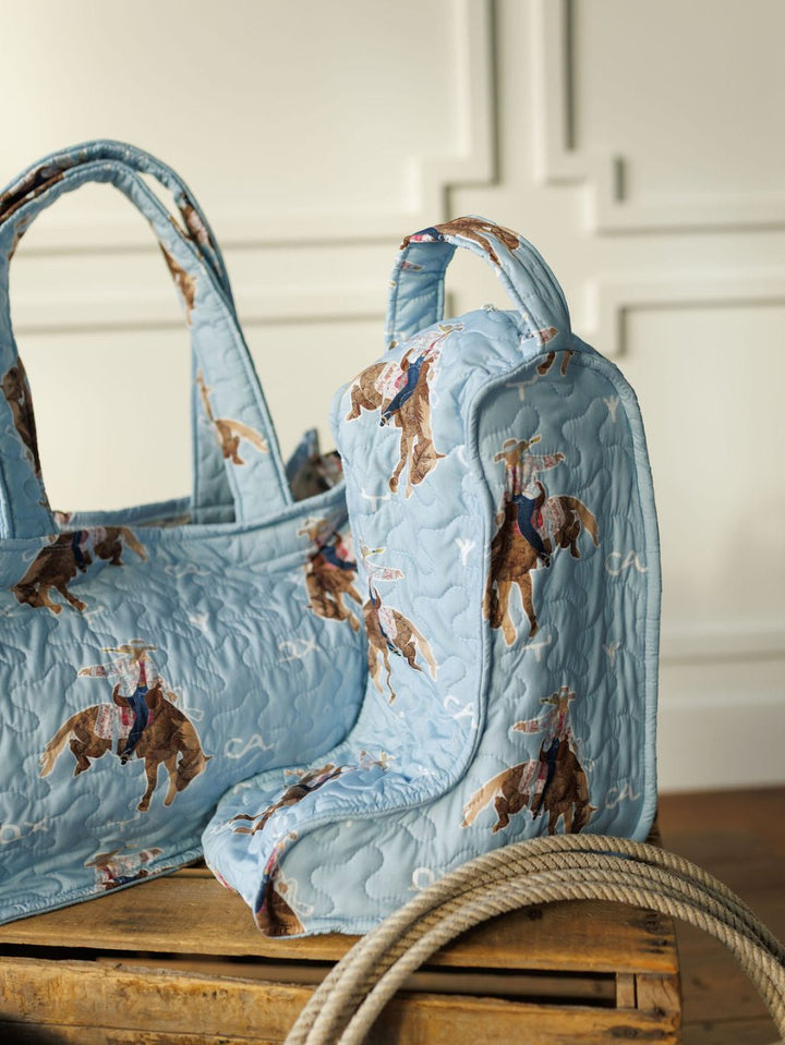 The Brumby Sky Collection quilted bag set and matching boot bag by Savannah Sevens western life&style, featuring a cowboy on a bucking bronco, rest on a wooden crate with a coiled rope against white paneled walls. By Ida Mae. 