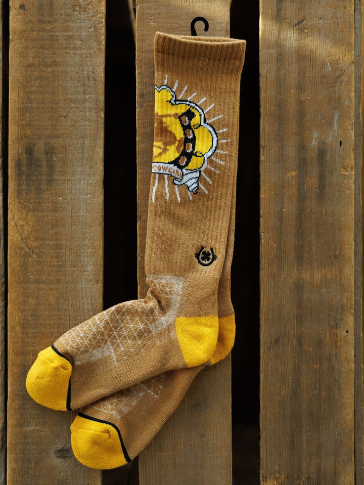 The Bucking Cowgirl Crew Socks by Savannah Sevens western life{&}style are brown and yellow with a cartoon cow skull graphic, yellow toes and heels, and a white grid on the foot, shown hanging on a wooden slatted background.