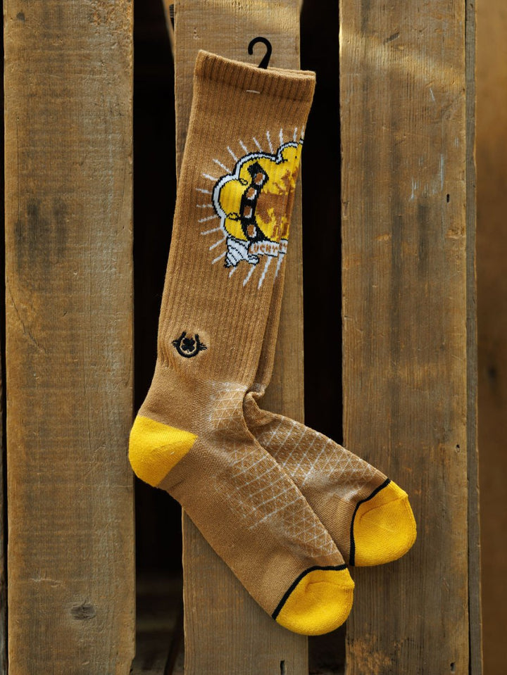 Bucking Cowgirl Crew Socks by Savannah Sevens western life{&}style hang on a wooden plank. These tan knee-high socks with yellow toes and heels feature a cartoon honey bee with a dripping honeycomb and a small ankle logo.