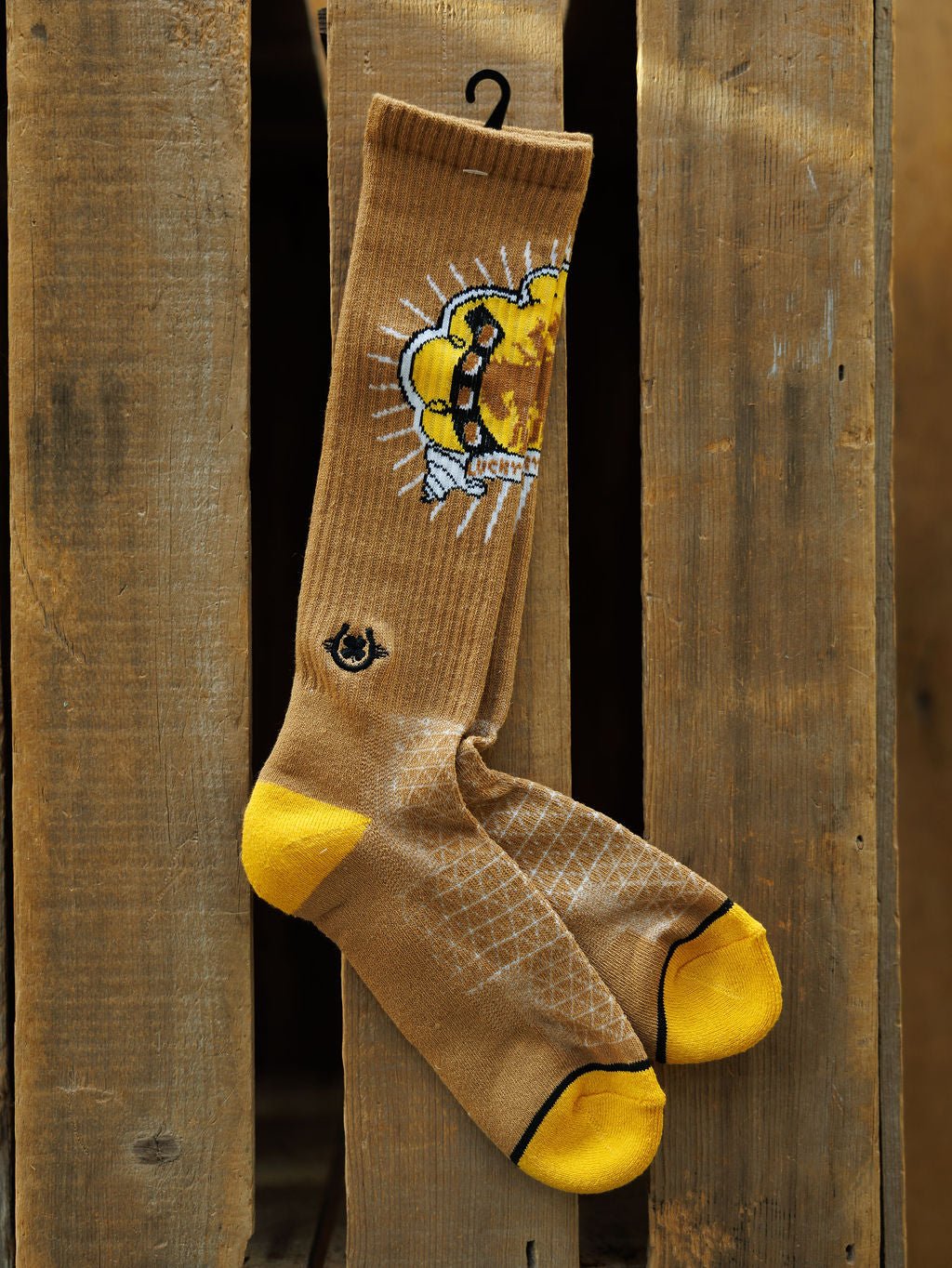 Bucking Cowgirl Crew Socks by Savannah Sevens western life{&}style hang on a wooden plank. These tan knee-high socks with yellow toes and heels feature a cartoon honey bee with a dripping honeycomb and a small ankle logo.