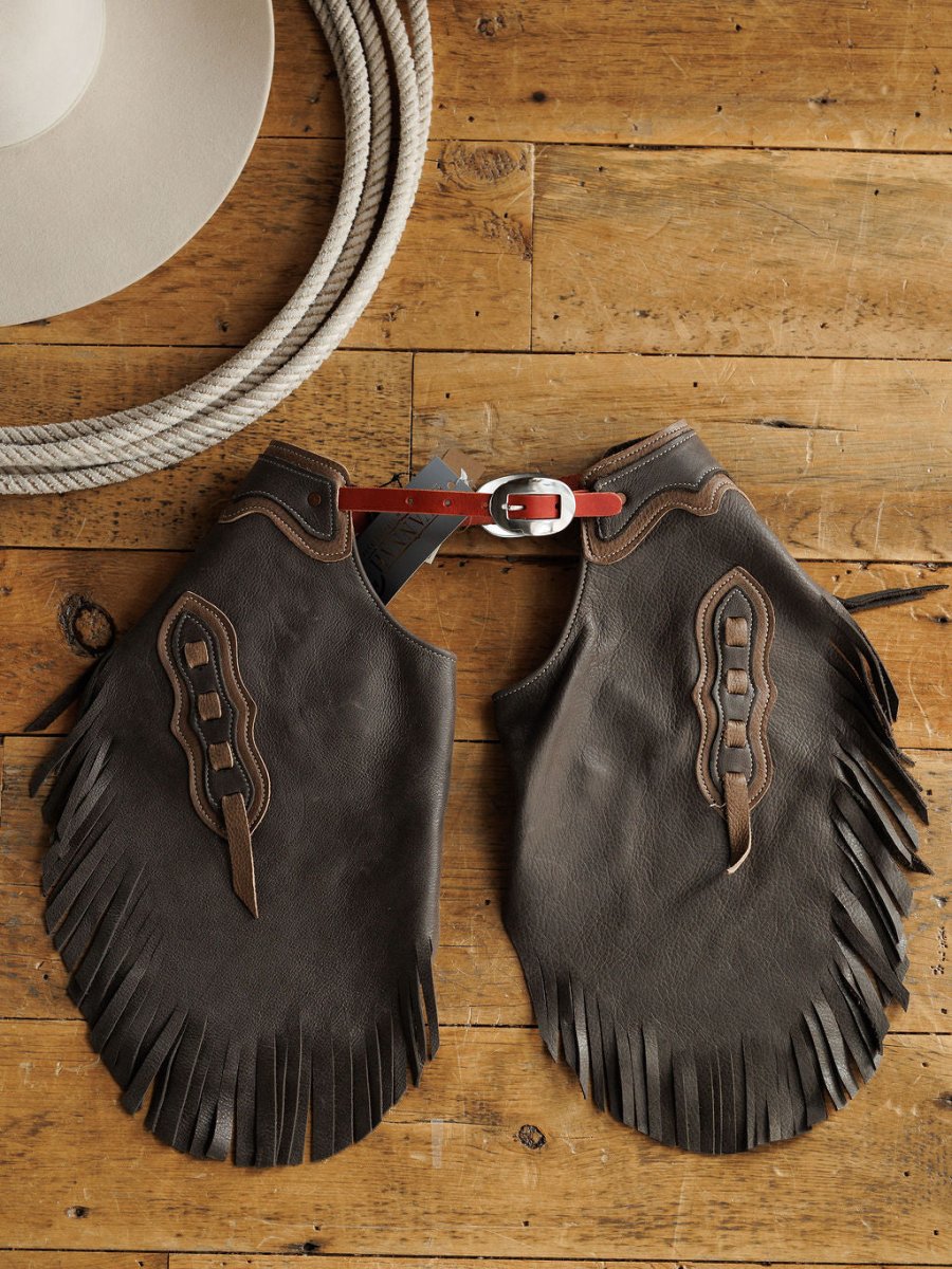 The Buffalo Chinks {Youth} by Savannah Sevens western life{&}style, featuring brown handmade leather with fringe and decorative stitching, are displayed on a wooden surface beside a coiled rope and a white cowboy hat.