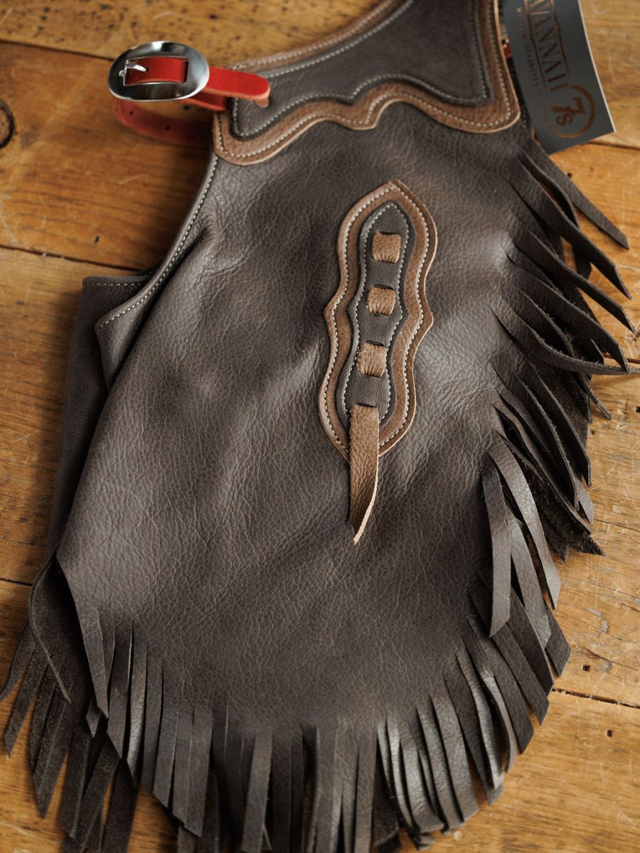 The Buffalo Chinks {Youth} from Savannah Sevens western life{&}style are brown handmade leather chinks with fringe, decorative stitching, and a red belt buckle, shown on wood with a brand tag attached.