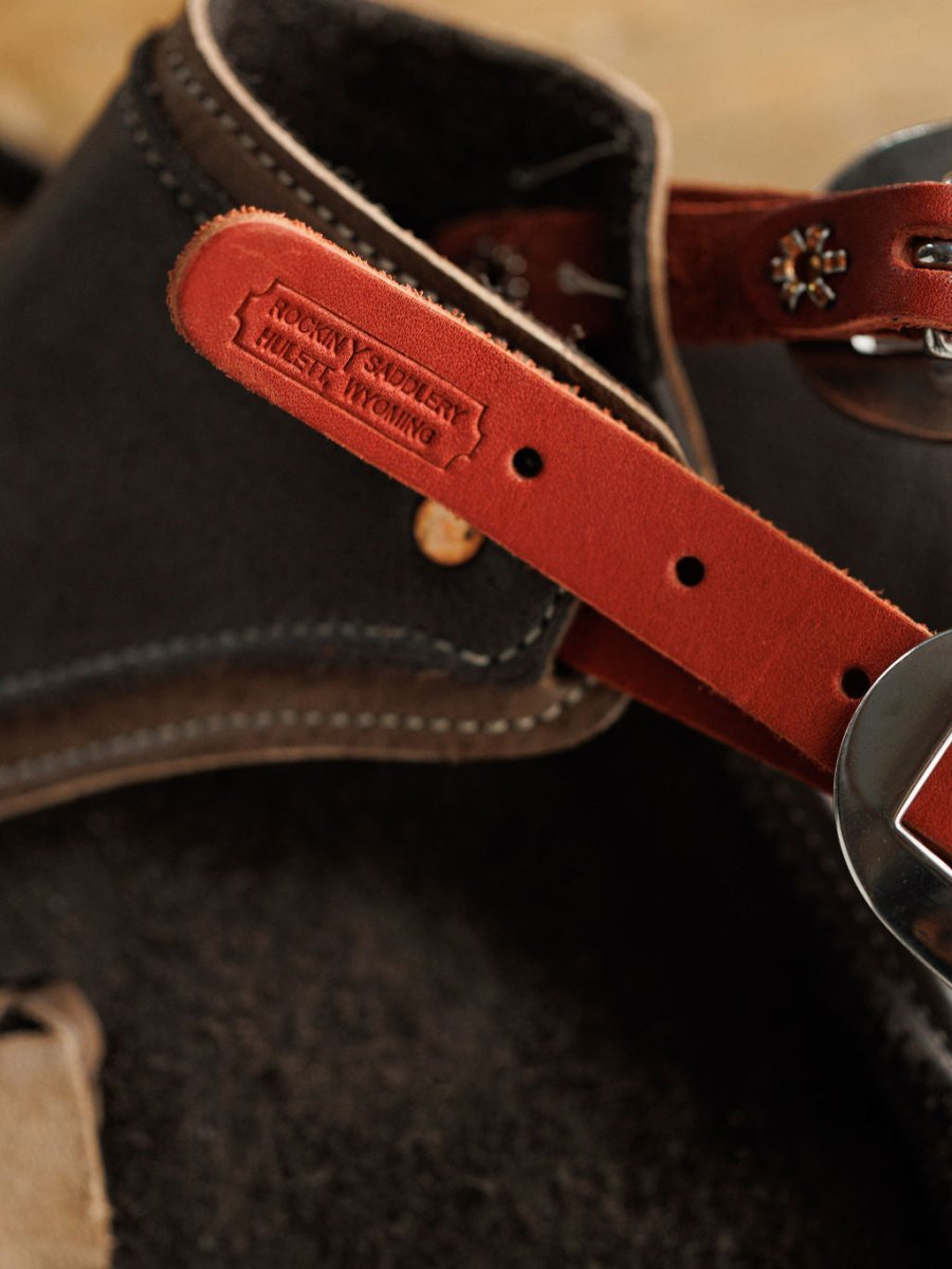 A close-up of Buffalo Chinks {Youth} by Savannah Sevens western life{&}style, showing a brown and red leather strap with detailed stitching, metal buckle, and embossed Pony Showheel Horse Wyoming, attached to dark brown cowboy chaps.