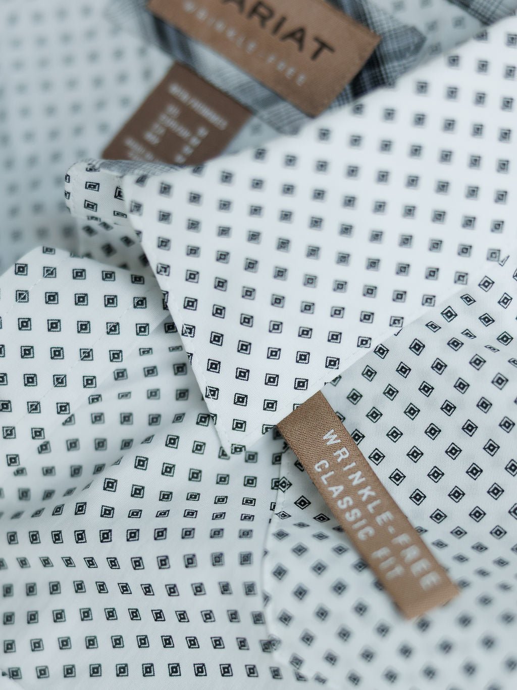 Close-up of the Burke Ariat Men's Southwest Pearl Snap from Savannah Sevens western life{&}style, featuring a small black square pattern, snap buttons, and brown tags reading Wrinkle Free Classic Fit and Ariat. Fabric looks crisp and neatly folded.