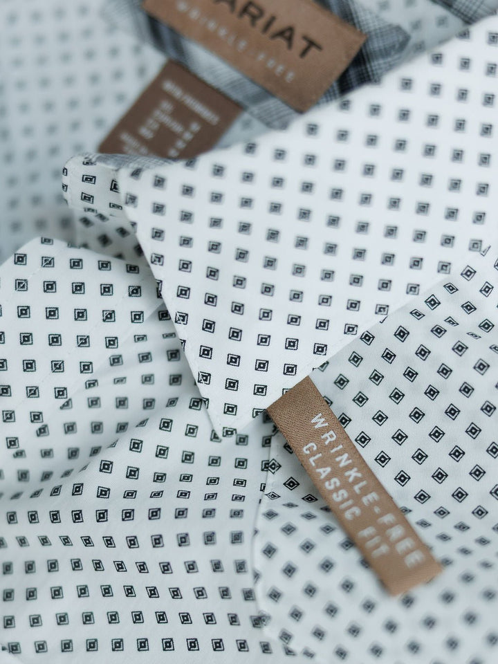 Close-up of the Burke Ariat Men's Southwest Pearl Snap from Savannah Sevens western life{&}style, featuring a small black square pattern, snap buttons, and brown tags reading Wrinkle Free Classic Fit and Ariat. Fabric looks crisp and neatly folded.