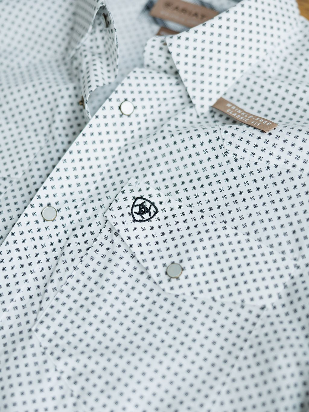 A close-up of the Burke Ariat Men's Southwest Pearl Snap by Savannah Sevens western life{&}style, showing a white shirt with a small black southwest pattern, silver snaps, an embroidered logo on the pocket, and a brown collar label.