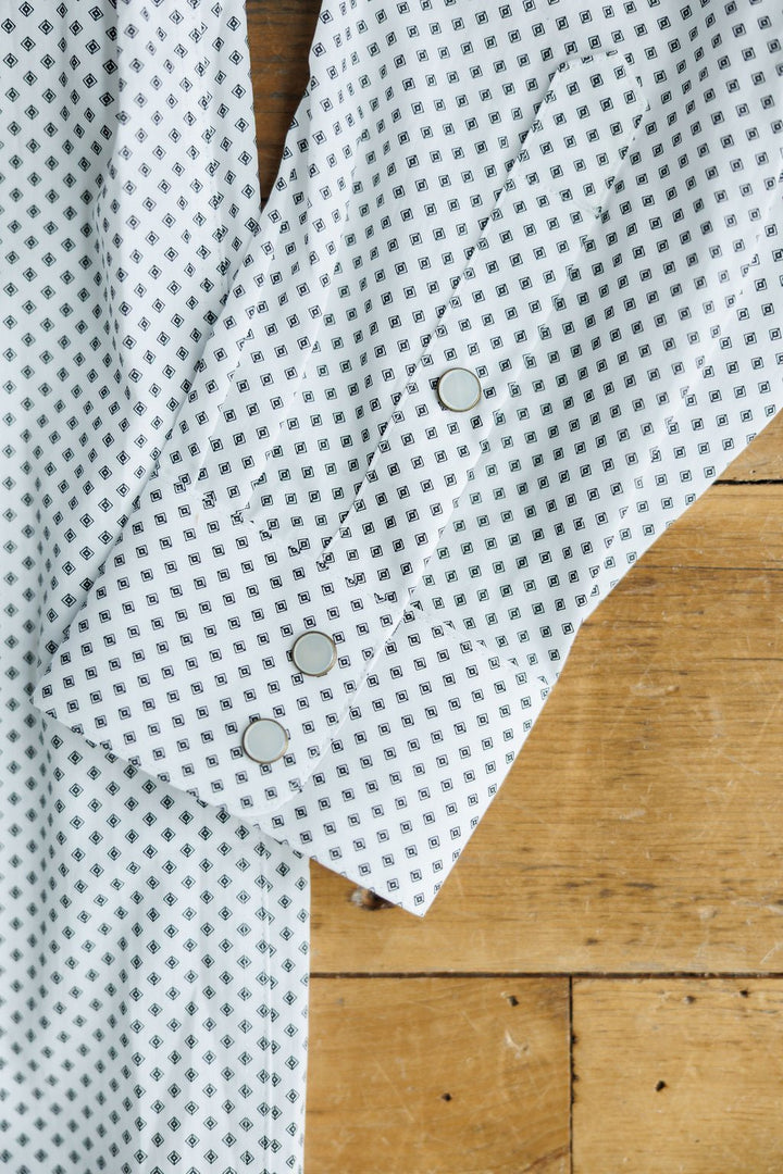A close-up of the Burke Ariat Men's Southwest Pearl Snap shirt from Savannah Sevens western life{&}style—a white shirt with a black geometric pattern and silver snap buttons, displayed on a wooden surface.
