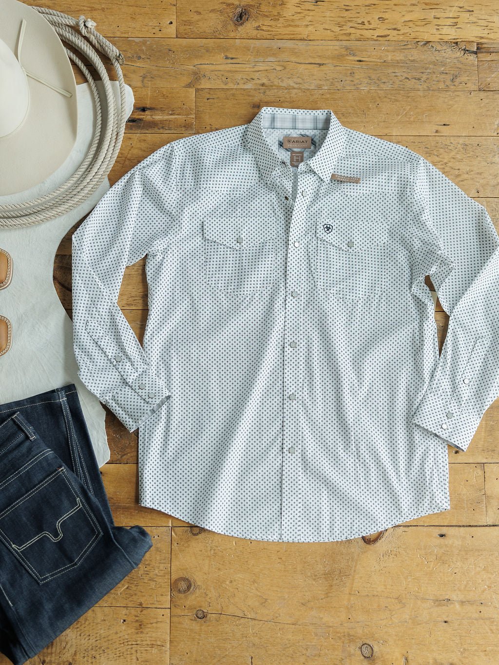 The Burke Ariat Men's Southwest Pearl Snap by Savannah Sevens western life{&}style is laid flat on a wooden floor beside dark jeans, a white cowboy hat, coiled rope, and leather chaps.