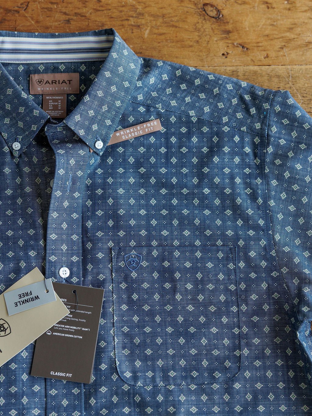 The Burleson Ariat Men's Southwest Button Up by Savannah Sevens western life{&}style features a blue long sleeve design with a subtle white geometric pattern. Displayed with “wrinkle free” tags, it offers versatile southwest style.