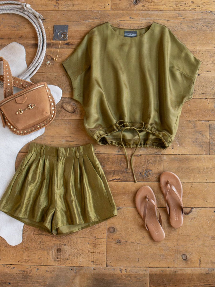 A flat lay featuring the Savannah Sevens western life{&}style Caddo Olive Satin Set—short-sleeve top and high-waisted shorts—styled with tan flip-flops, a studded tan crossbody bag, and a coiled rope on a wooden floor.