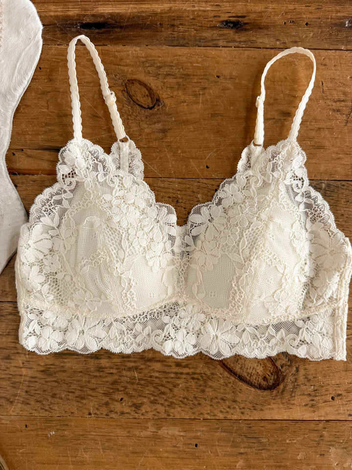 The Cairo Cream Lace Bralette by Savannah Sevens western life{&}style features delicate floral lace and adjustable straps, shown laid flat on a rustic wooden surface.