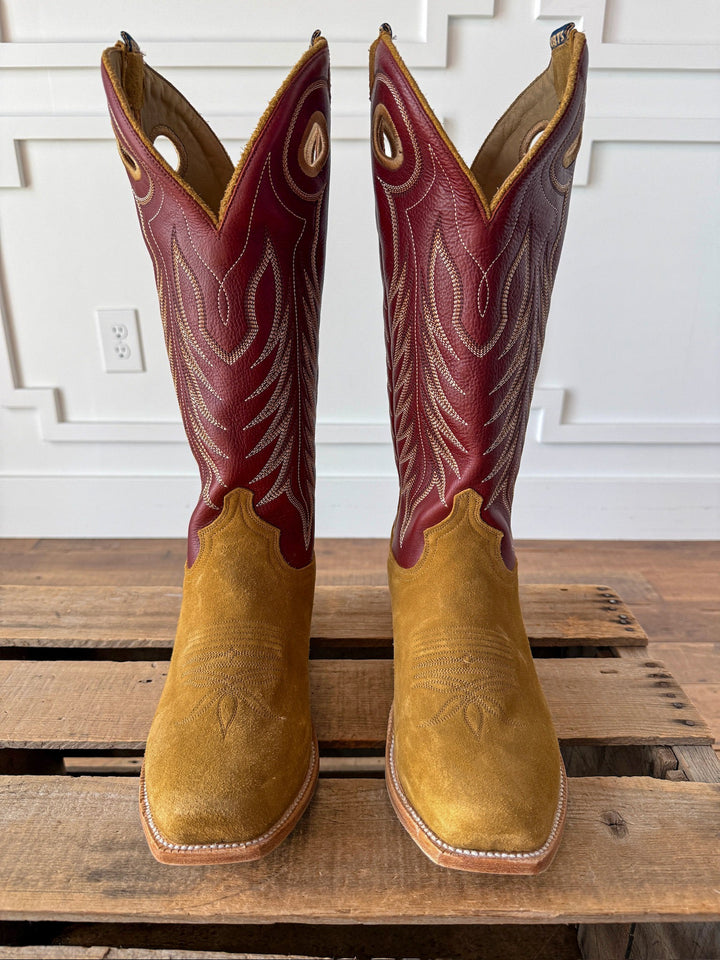 A pair of Caldwell Tan Men’s Western Leather Boots by Savannah Sevens western life{&}style, featuring tan suede feet and red leather shafts with white decorative stitching, displayed on a wooden pallet against a white paneled wall. This men’s leather boot showcases a classic cutter toe design. Boots by HYER. 