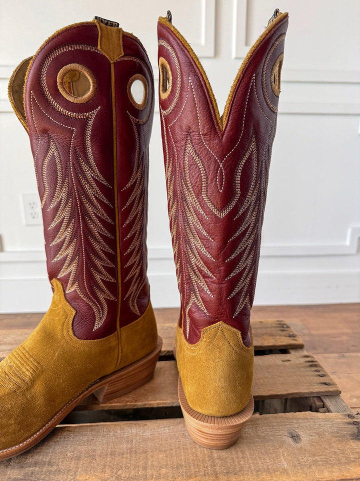 A pair of Caldwell Tan Men’s Western Leather Boots by Savannah Sevens western life{&}style, featuring a yellow suede foot, maroon leather shaft with intricate white stitching, round pull holes, and a classic cutter toe boot design, displayed on a wooden surface. Boots by HYER. 