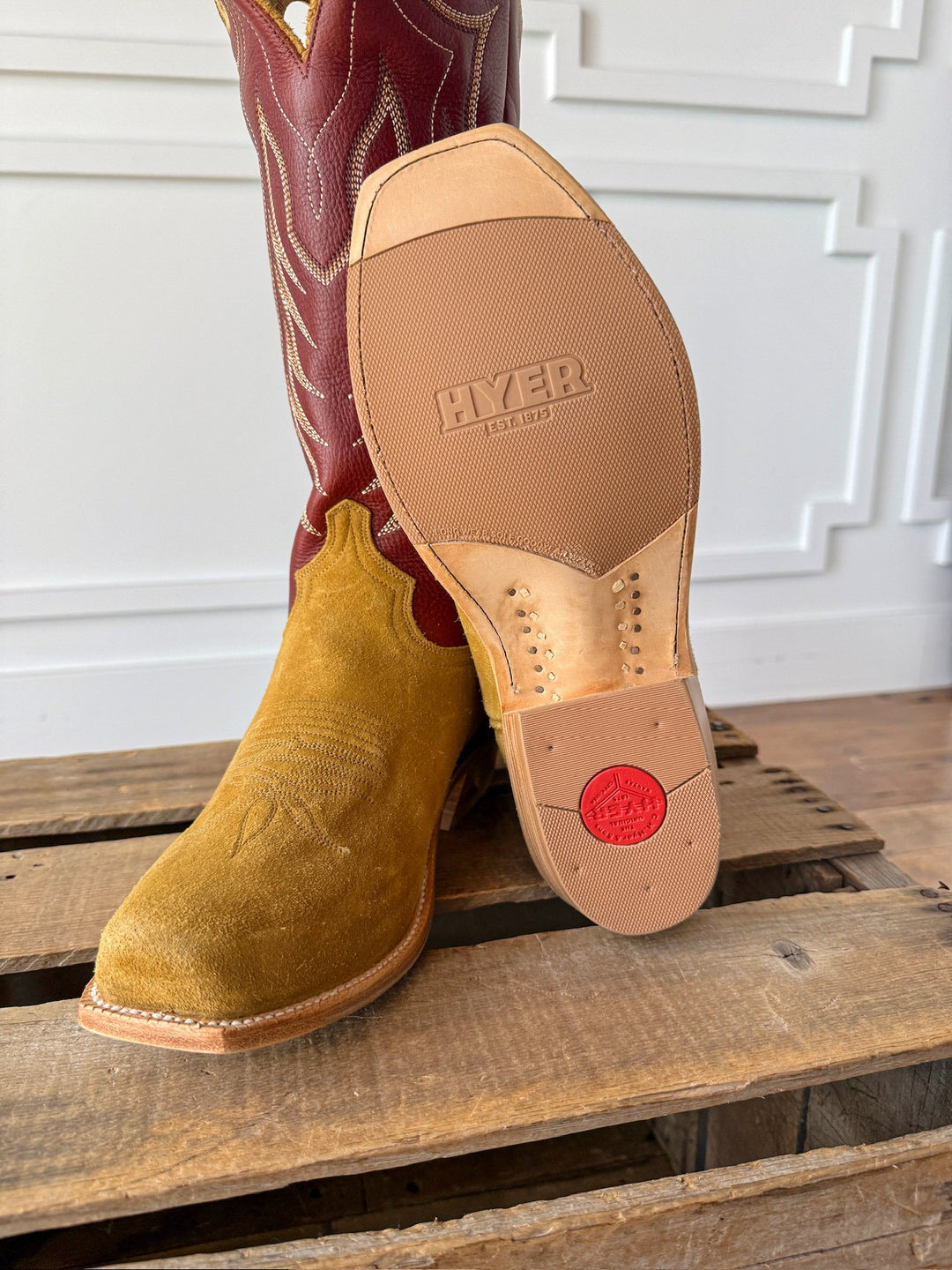 A close-up of the Caldwell Tan Men’s Western Leather Boot by Savannah Sevens western life{&}style rests on a wooden surface, highlighting its tan suede upper, red and tan shaft adorned with intricate stitching, and a sole stamped with HYER EST. 1875 beside a distinctive red circle near the heel.