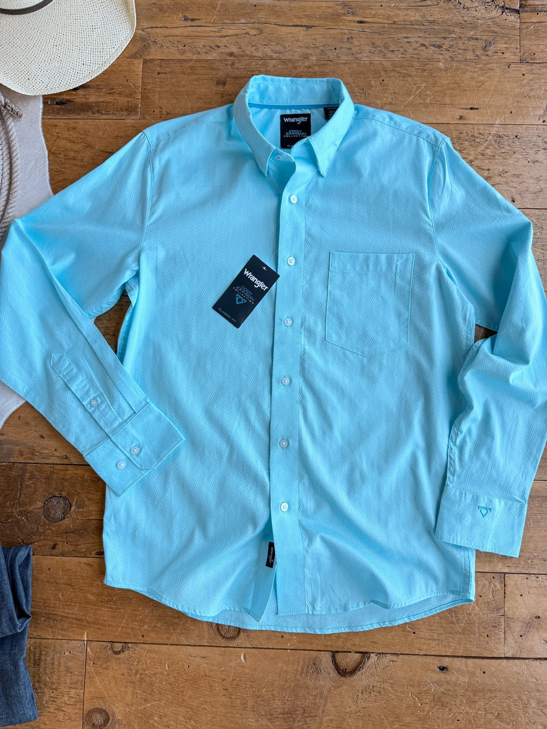 A light blue "The Caldwell" men's long sleeve button up shirt by Savannah Sevens western life{&}style, 100% cotton with tags, is laid out on a wooden floor. Edges show part of a hat, sweater, and jeans.