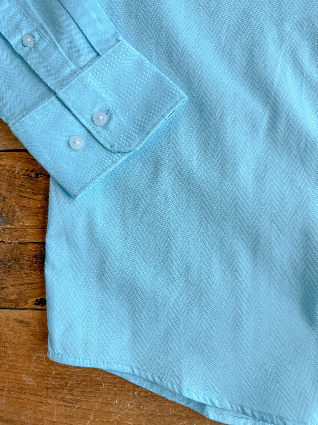 A close-up of The Caldwell men's long sleeve button up shirt by Savannah Sevens western life&style, in light blue with a subtle herringbone pattern, made from 100% cotton. The shirt is laid flat on wood, displaying the cuff and body.