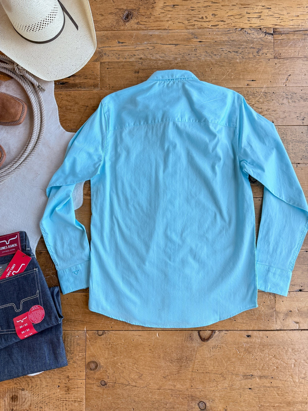 The Caldwell Men's shirt by Savannah Sevens western life&style, in light blue 100% cotton, is laid flat with the back up on a wooden floor, surrounded by a white cowboy hat, brown boots, beige vest, and folded blue jeans.