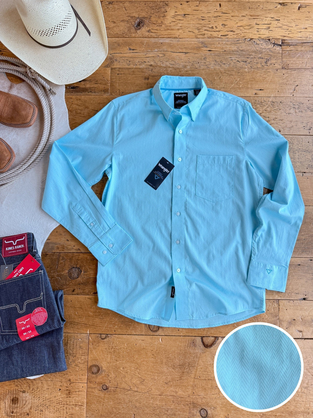 A light blue Caldwell men’s long sleeve button up from Savannah Sevens western lifestyle is laid flat on a wooden floor beside folded jeans, brown boots, and a cowboy hat. An inset showcases its subtle textured pattern and 100% cotton fabric.