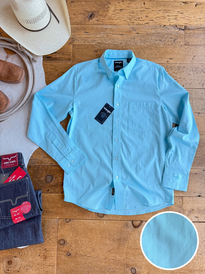 A light blue Caldwell men’s long sleeve button up from Savannah Sevens western lifestyle is laid flat on a wooden floor beside folded jeans, brown boots, and a cowboy hat. An inset showcases its subtle textured pattern and 100% cotton fabric.