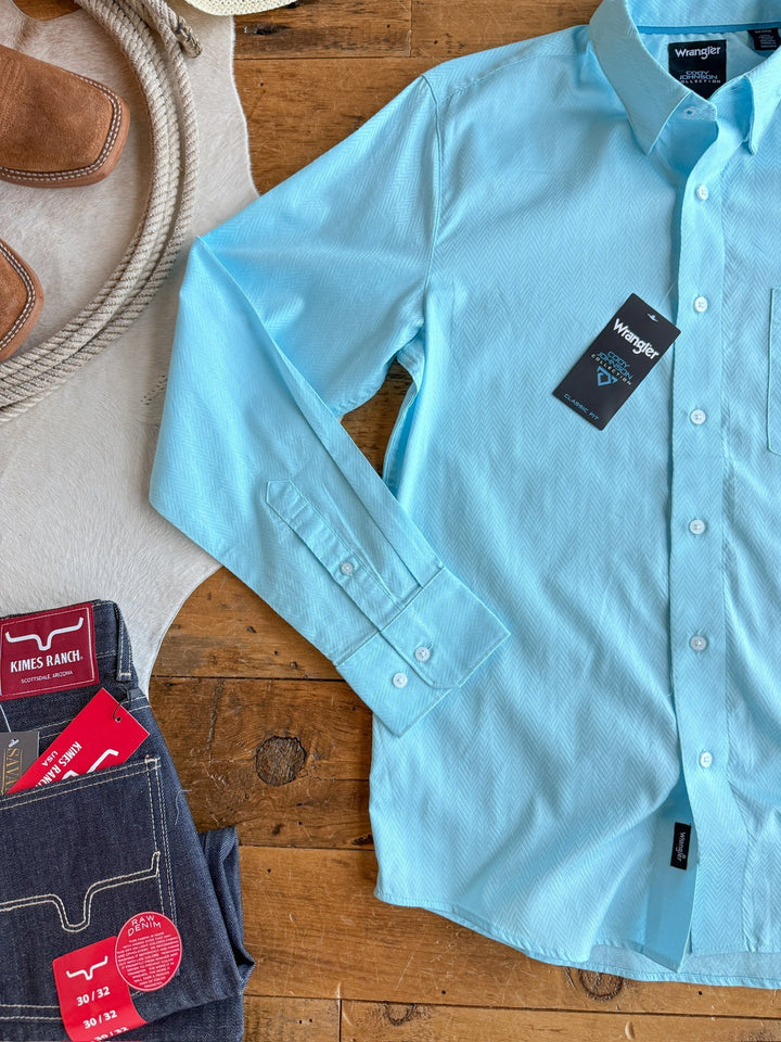 A light blue 100% cotton "The Caldwell" men's long sleeve button-up shirt by Savannah Sevens western life{&}style is displayed with dark blue jeans, a coiled rope, and a cowboy hat on a wooden surface.