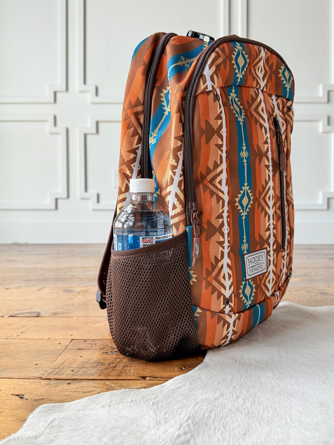 The Calexico Backpack by Savannah Sevens western life{&}style, featuring geometric Aztec designs in brown, orange, and blue, rests on a wooden floor with a water bottle in its mesh pocket. White paneled walls and a white rug are seen in the background. By Hooey. 