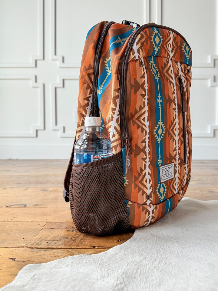 The Calexico Backpack by Savannah Sevens western life{&}style, featuring geometric Aztec designs in brown, orange, and blue, rests on a wooden floor with a water bottle in its mesh pocket. White paneled walls and a white rug are seen in the background. By Hooey. 