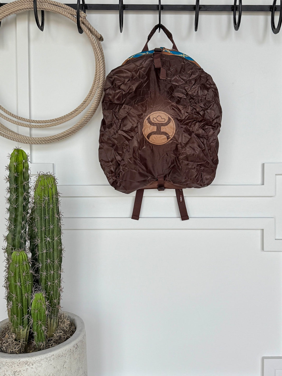 A brown Calexico Backpack by Savannah Sevens western life{&}style with a light brown circular logo hangs on a black hook beside a coiled rope; below, a tall potted cactus stands against a white paneled wall. By Hooey. 